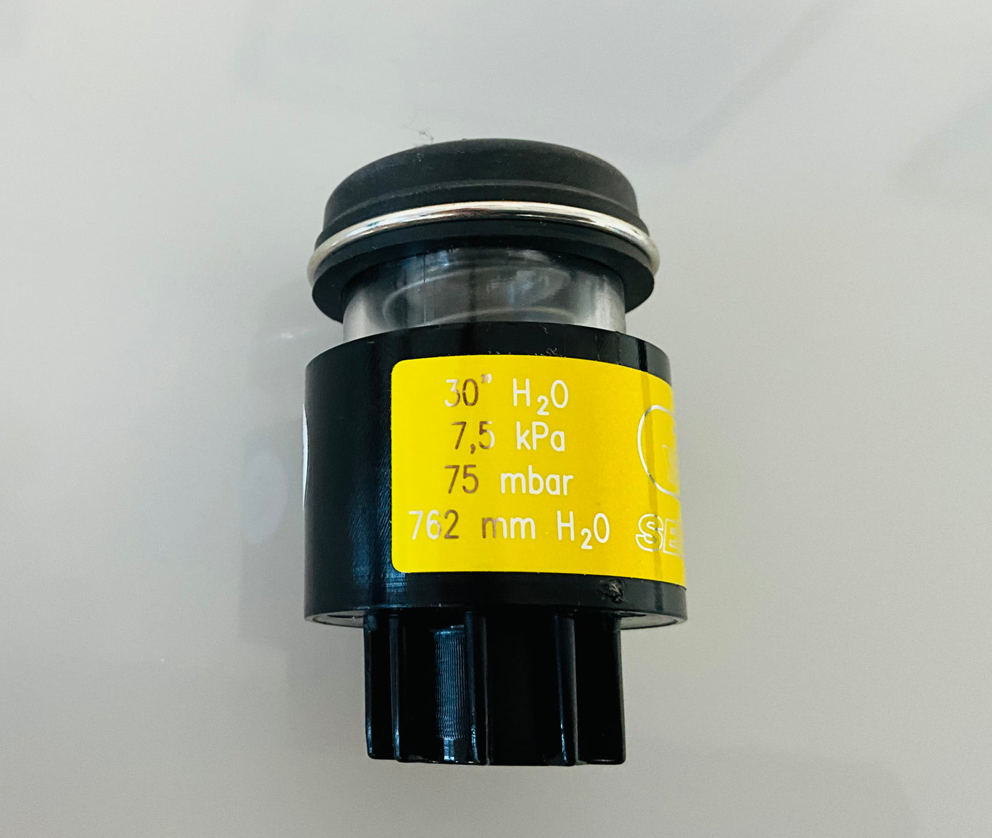 A small black cylindrical optical indicator with press button top, with a yellow label that reads '30 H2O', '7,5 kPa', '75 mbar', '762 mm H2O'. Shown on a reflective white surface.
