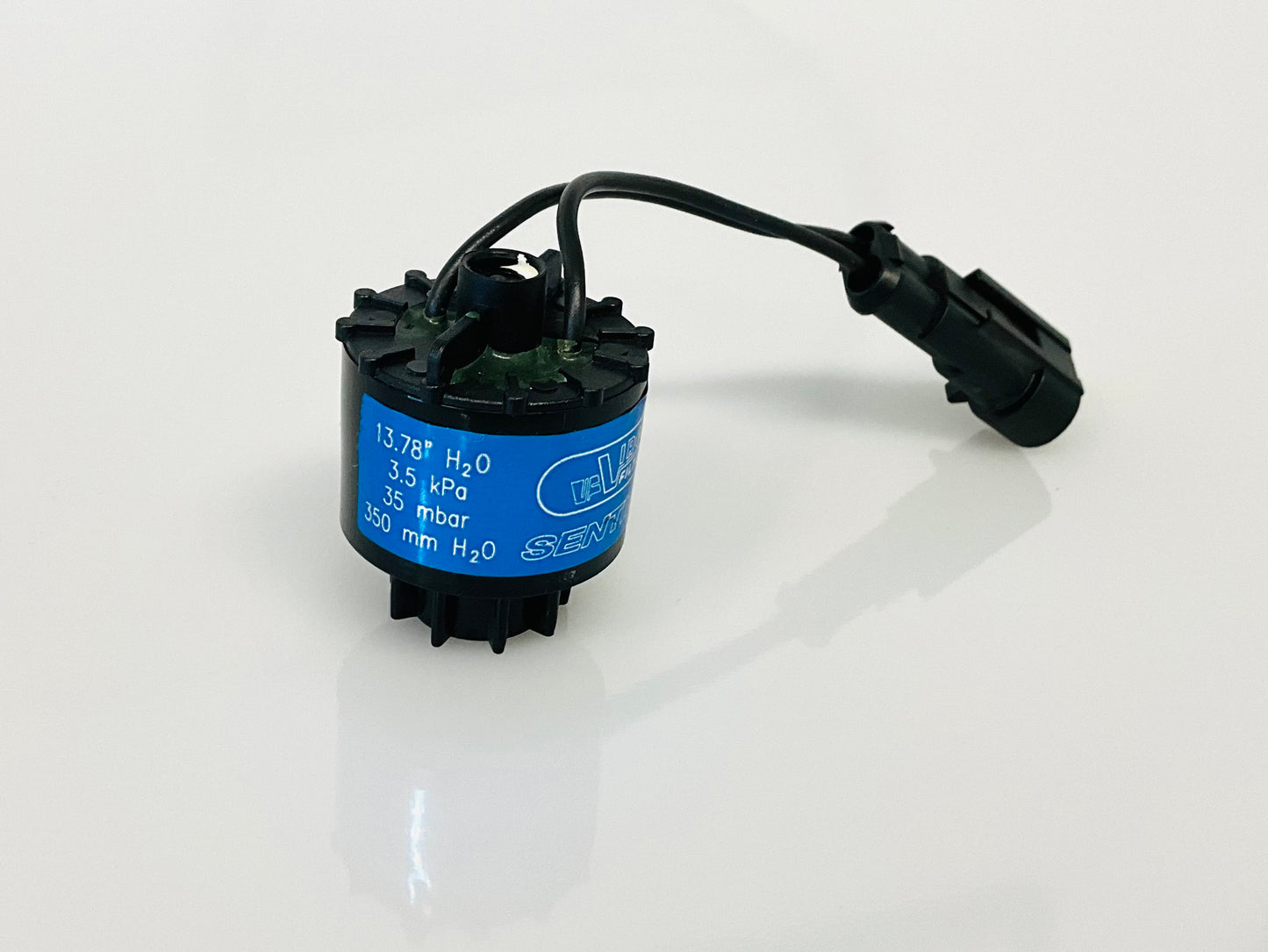 A black electrical indicator with a cable connection and blue label, that reads '13.78" h2O', '305 kPa', '35 mbar', '350 mm h2O', shown on a reflective white surface.