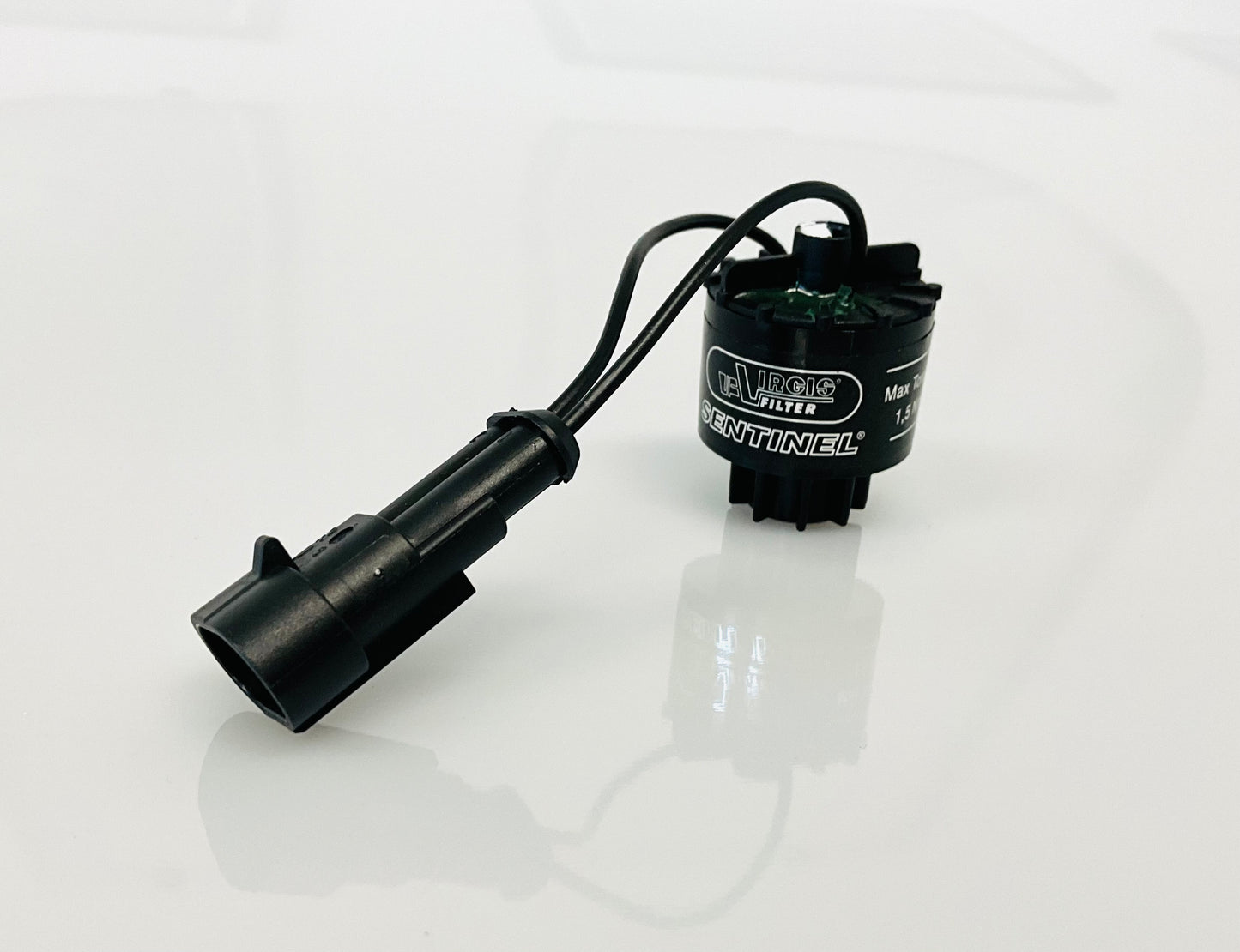 A black electrical indicator with a cable connection and a label, that reads 'Virgis Filter' and 'Sentinel'. Shown on a reflective white surface.