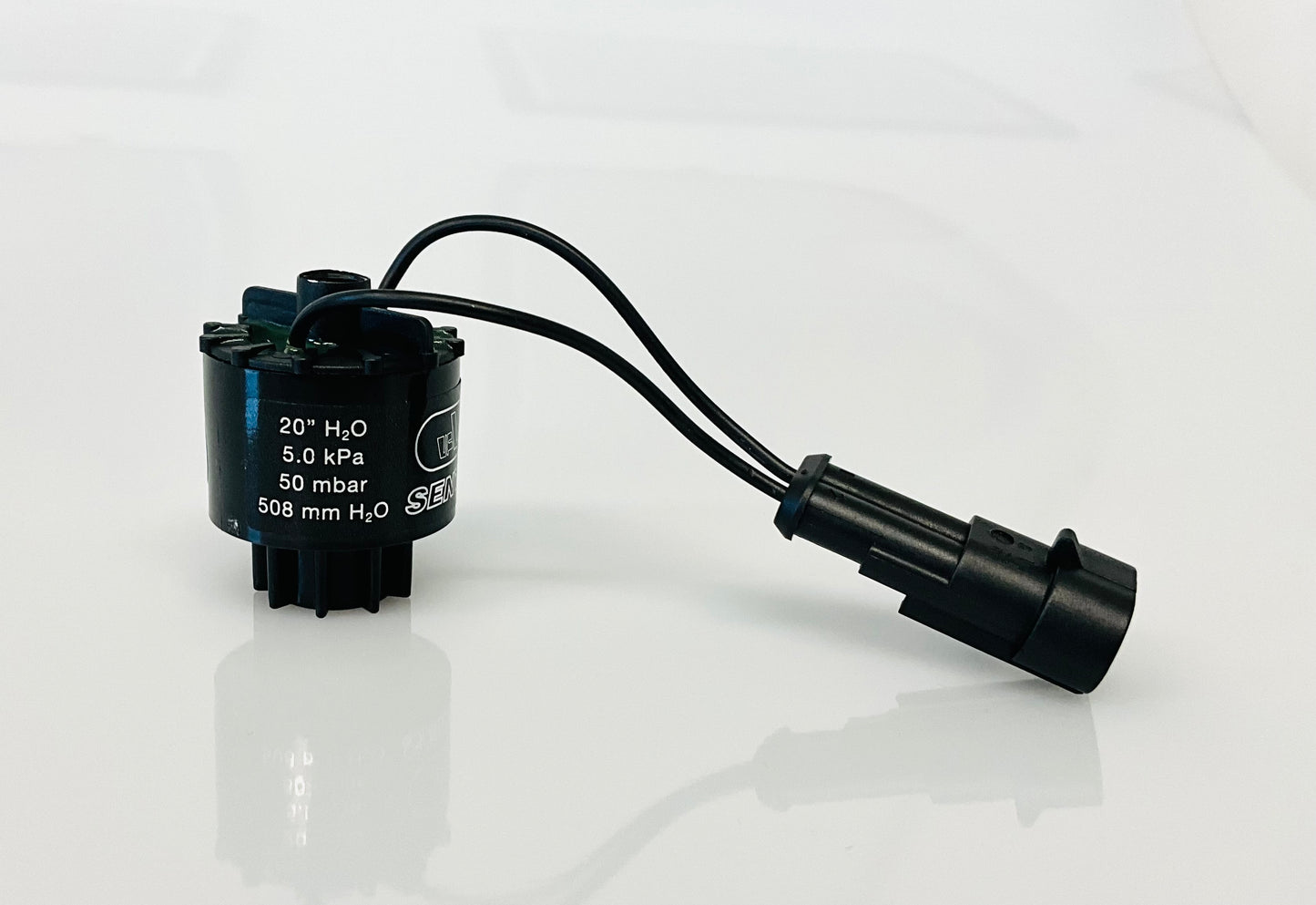 A black electrical indicator with a cable connection and a label, that reads '20" H2O', '5.0 kPa', '50 mbar', 508 mm h2O', shown on a reflective white surface.