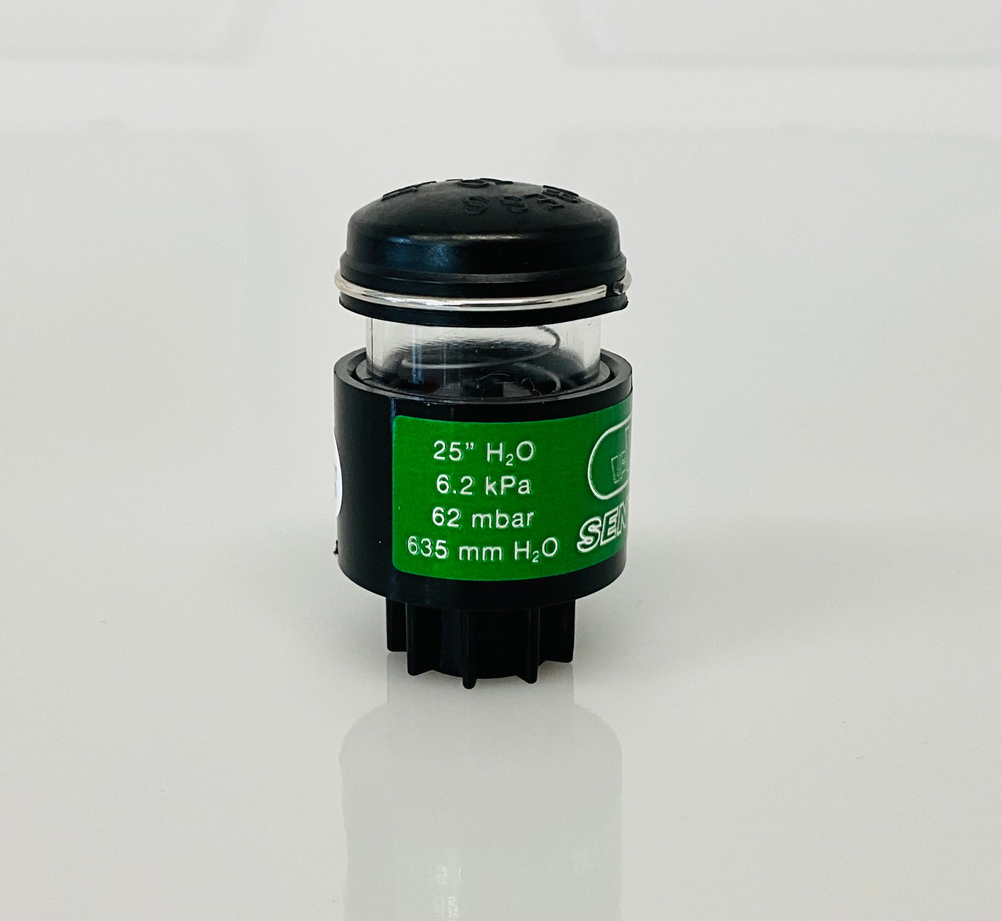 A small black cylindrical optical indicator with press button top, with a green label reading '25" H2O 6.2 kPa 62 mbar 635 mm H2O', photographed on a white background.
